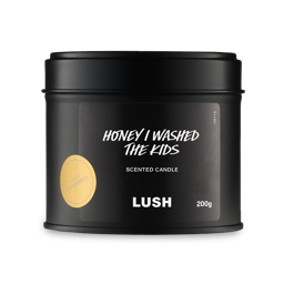 A short cylindrical metallic black tin with a metallic black lid and a black label that has Lush Honey I Washed The Kids Single Wick Candle written in bright white writing and Community written in bright white writing inside a bright gold circle on it, on a white background. 