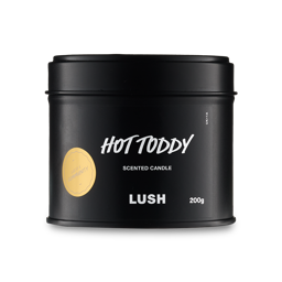 A short cylindrical metallic black tin with a metallic black lid and a black label that has Lush Hot Toddy Single Wick Candle written in bright white writing and Community written in bright white writing inside a bright gold circle on it, on a white background. 