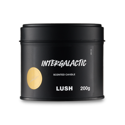A short cylindrical metallic black tin with a metallic black lid and a black label that has Lush Intergalactic Single Wick Candle written in bright white writing and Community written in bright white writing inside a bright gold circle on it, on a white background. 