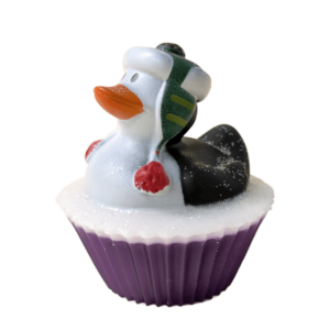 A short cylindrical bright white cupcake shaped block of soap on a sparkly dark purple cupcake liner that has a black and white rubber duck with a bright orange beak wearing a dark green hat with some bright red pompoms sat on top of it, on a white background.