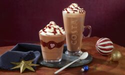A tall and wide cylindrical clear glass full of some dark brown liquid and some bright white whipped cream that has Costa and a picture of a coffee Bean engraved on it next to a tall cylindrical clear glass cup full of some dark brown liquid and some bright white whipped cream that has Costa engraved on it on a large circular slate serving board next to a small gold star tree decoration and a dark blue Napkin, on a dark red background.