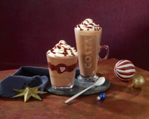 A tall and wide cylindrical clear glass full of some dark brown liquid and some bright white whipped cream that has Costa and a picture of a coffee Bean engraved on it next to a tall cylindrical clear glass cup full of some dark brown liquid and some bright white whipped cream that has Costa engraved on it on a large circular slate serving board next to a small gold star tree decoration and a dark blue Napkin, on a dark red background.