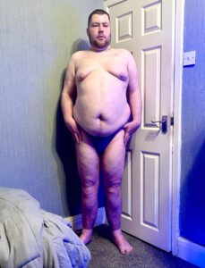 A tall bearded man with his hands on his hips wearing a dark blue tanga brief with a black waistband that has Kiniki written in bright white writing on it stood against a white wooden door.