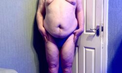 A tall bearded man with his hands on his hips wearing a dark blue tanga brief with a black waistband that has Kiniki written in bright white writing on it stood against a white wooden door.