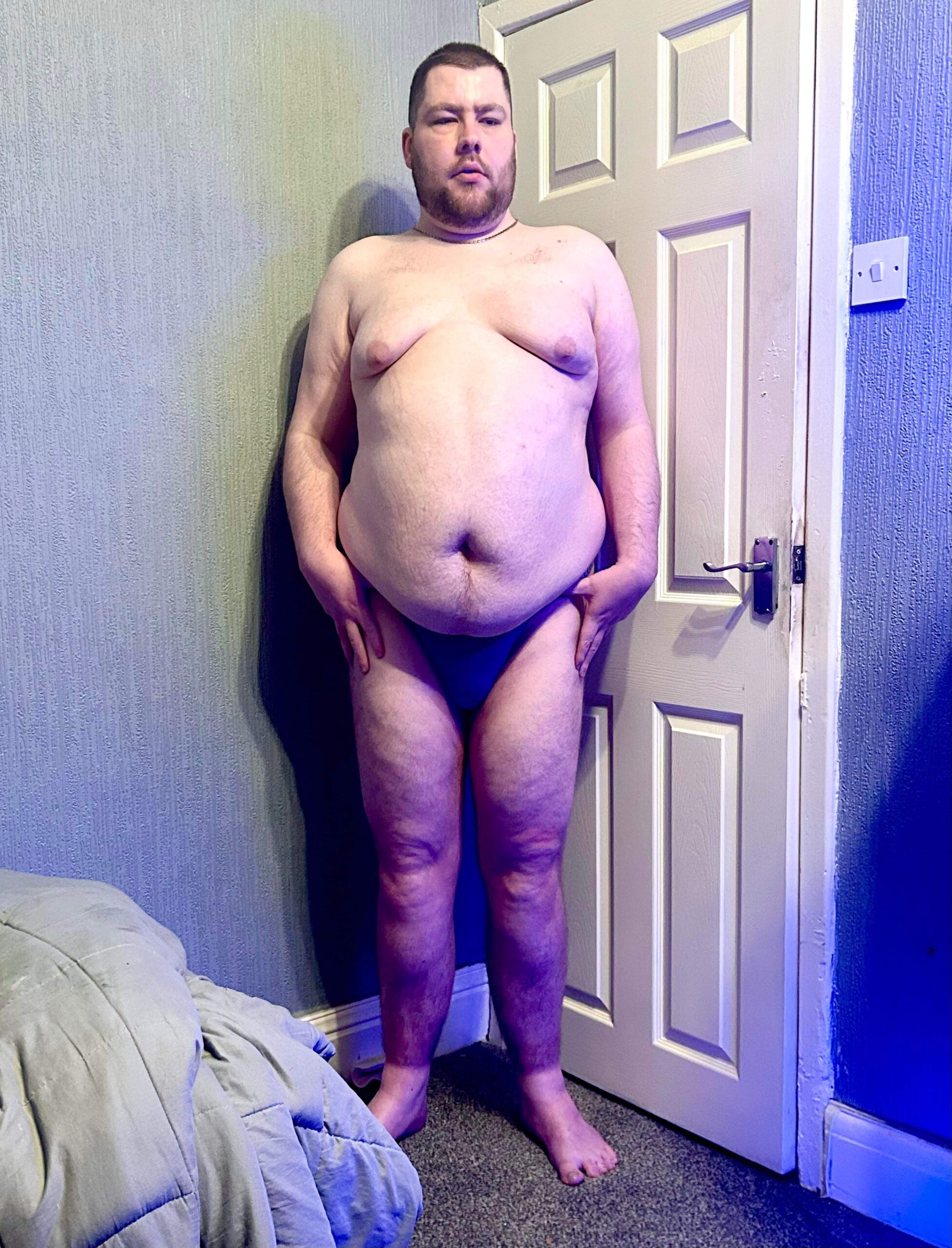 A tall bearded man with his hands on his hips wearing a dark blue tanga brief with a black waistband that has Kiniki written in bright white writing on it stood against a white wooden door.