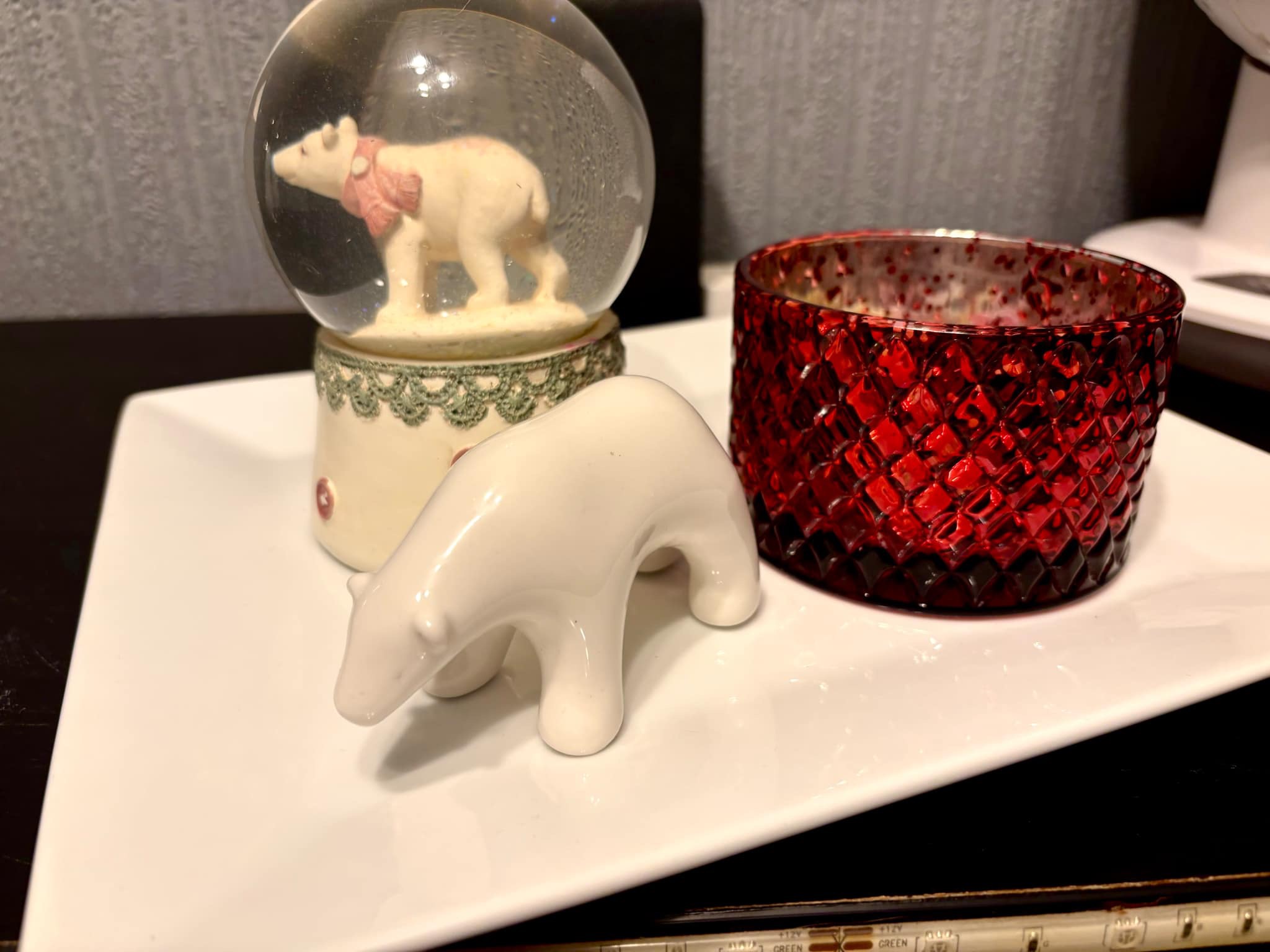A large bright white spherical snow globe that has a polar bear stood on all four paws inside it next to a small white polar bear that’s stood on all four paws on top of a large rectangular white plate and is surrounded by some clear white strip lights, on a black wooden cabinet.