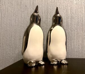 Two tall silver ceramic Penguin ornaments that both have white bellies, on a black wooden cabinet.