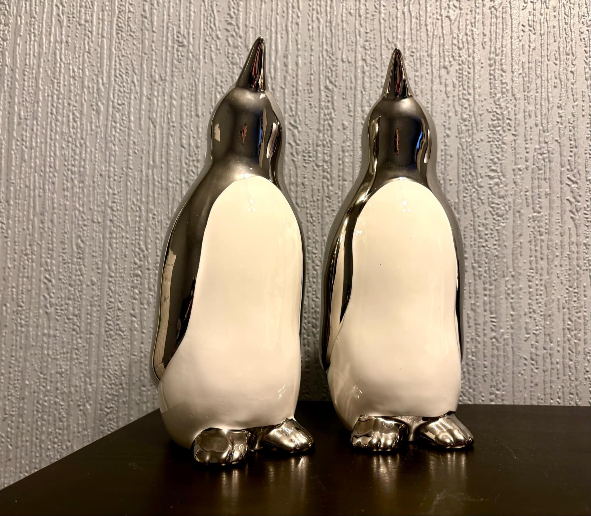 Two tall silver ceramic Penguin ornaments that both have white bellies, on a black wooden cabinet.