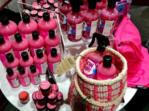 Several tall cylindrical clear plastic bottles full of some bright pink shower gel next to a pyramid shaped pile of some short cylindrical plastic tubs full of some bright pink liquid, a large cylindrical black woven basket and a large rectangular bright pink makeup bag on a bright white circular table.