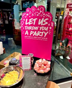 A large spherical black metal bowl full of some bright yellow powder next to a large circular black plate that has a pile of some bright pink, orange, yellow and white Santa Clause shaped bath Bombs on it on a light wooden table in front of a large rectangular bright pink sign that has Let Us Throw You A Party written in bright white writing and some pictures of some Balloons on it, on a bright background.