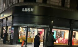 A large rectangular brightly lit dark brown brick store front with a large rectangular black board over its door that has Lush written in bold white writing on it on a dimly lit dark cobbled road..