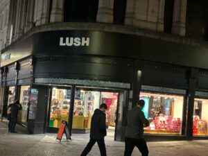 A large rectangular brightly lit dark brown brick store front with a large rectangular black board over its door that has Lush written in bold white writing on it on a dimly lit dark cobbled road..
