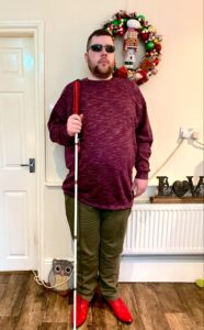 A tall bearded man wearing a dark burgundy jumper, a wine red V-Neck t-shirt, a pair of khaki green chino trousers and a pair of shiny red shoes holding a bright white cane with a red leather handle stood against a cream wall.