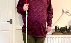 A tall bearded man wearing a dark burgundy jumper, a wine red V-Neck t-shirt, a pair of khaki green chino trousers and a pair of shiny red shoes holding a bright white cane with a red leather handle stood against a cream wall.