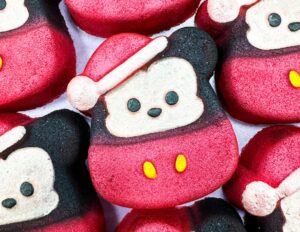 A pile of some large Micky Mouse Head shaped bath bombs that all have a bright red Santa’s hat, a bright red coat with some bright white buttons, and a bright white face with some black eyes, a black nose, and some black ears engraved on them, on a white background.