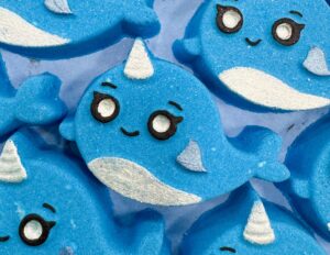 A pile of some medium bright blue Narwal shaped bath bombs that all have a bright white horn, a bright white belly, and a cartoon Face with some big eyes and a small smile engraved on them, on a light blue background.