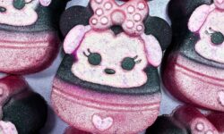 A pile of some bright white Minnie Mouse Head shaped Bath Bombs that all have a bright pink and white polka dot bow, a small bright pink heart, and a bright white face with some black eyes, a black nose and some bright pink ears on them, on a white background.