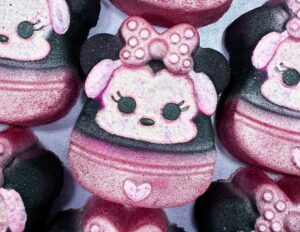 A pile of some bright white Minnie Mouse Head shaped Bath Bombs that all have a bright pink and white polka dot bow, a small bright pink heart, and a bright white face with some black eyes, a black nose and some bright pink ears on them, on a white background.