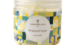 A short cylindrical clear Plastic Tub full of some golden yellow whipped soap with a clear Plastic Lid and a bright blue, green, white and yellow multicoloured label that has The Yorkshire Soap Limoncello Whipped Soap written in black writing inside a white circle and some pictures of some bright yellow lemons on it, on a white background.