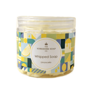 A short cylindrical clear Plastic Tub full of some golden yellow whipped soap with a clear Plastic Lid and a bright blue, green, white and yellow multicoloured label that has The Yorkshire Soap Limoncello Whipped Soap written in black writing inside a white circle and some pictures of some bright yellow lemons on it, on a white background.