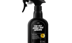 A tall black plastic kitchen spray bottle that has Lush The Old Songs Are Waking Body Spray written in bright white writing on it, on a white background.