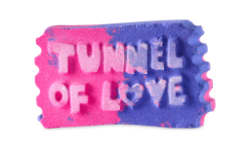 A medium gradient bright pink and purple ticket stub shaped bath bomb that has Tunnel Of Love written in bright white writing and a picture of a love heart engraved on it, on a white background.