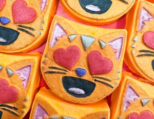 A pile of some bright orange cat’s head shaped bath bombs that all have some red love heart eyes, some small bright blue noses, some large black mouths, and some shiny dark purple ears on them, on a white background.