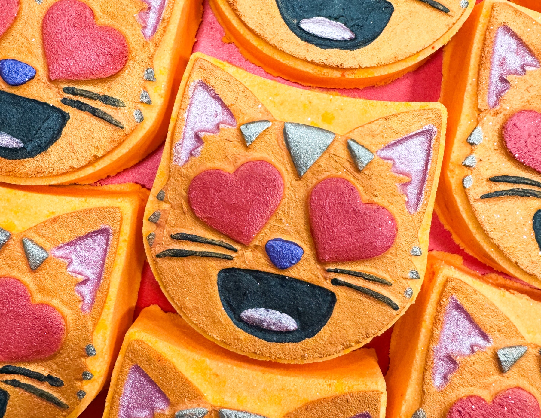 A pile of some bright orange cat’s head shaped bath bombs that all have some red love heart eyes, some small bright blue noses, some large black mouths, and some shiny dark purple ears on them, on a white background.