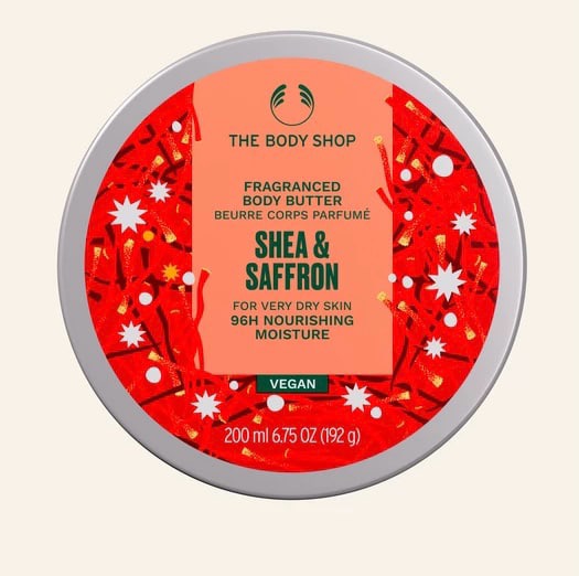 A short cylindrical dark green plastic tub with a dark red lid and a dark red label that has The Body Shop Shea & Saffron Body Butter written in dark green writing and some pictures of some bright white stars on it, on a white background.