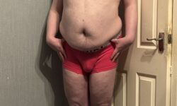 A tall bearded man with his hands on his hips wearing a pair of bright pink hipster briefs with a black waistband that has Kiniki written in bright white writing on it stood against a white wooden door.