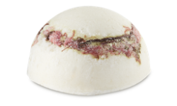 A medium half sphere shaped block of creamy white solid body scrub that has some pieces of granulated sugar running through it and some bright pink petals in the centre of it, on a white background.