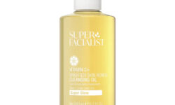 A tall cylindrical clear plastic bottle full of some amber coloured liquid with a clear plastic lid that has Super Facialist Vitamin C+ Brightening Cleansing Oil written in bright white writing on it, on a white background.