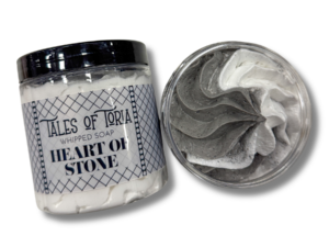 A tall cylindrical clear plastic tub full of some gradient bright grey and white whipped soap with a black plastic lid and a bright white label that has Tales of Toria Heart of Stone Whipped Soap written in bold black writing and some pictures of some black diamonds on it, on a white background.