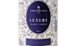 A tall bright white cardboard tube that has The Yorkshire Soap Company Afternoon Tea Glass Candle written in bright white writing inside a dark blue oval, a picture of a black crown, and some pictures of some bright white leaves on it, on a white background.