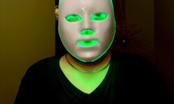 A tall bearded man wearing a charcoal grey v-neck t-shirt, a bright gold Chain and a bright white plastic LED Face Mask that’s covered in some green light stood against a light grey wall.