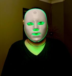 A tall bearded man wearing a charcoal grey v-neck t-shirt, a bright gold Chain and a bright white plastic LED Face Mask that’s covered in some green light stood against a light grey wall.