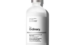 A tall cylindrical bright white glass bottle with a metallic silver lid that has The Ordinary Saccharomyces Ferment 30% Milky Toner written in bold black writing on it, on a white background.
