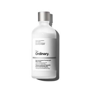 A tall cylindrical bright white glass bottle with a metallic silver lid that has The Ordinary Saccharomyces Ferment 30% Milky Toner written in bold black writing on it, on a white background.