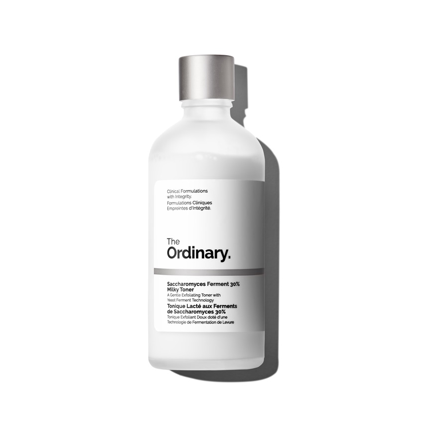 The Extraordinary Power Of A Hydrating Milky Toner | The Ordinary Saccharomyces Ferment 30% Milky Toner A tall cylindrical bright white glass bottle with a metallic silver lid that has The Ordinary Saccharomyces Ferment 30% Milky Toner written in bold black writing on it, on a white background.
