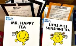 A large rectangular bright yellow cardboard box that has Bird & Blend Tea Company Little Miss Sunshine Gift Cube written in bold black writing and a picture of a bright yellow little miss sunshine that has a smiling face and some flowers in her hair on it next to a short cylindrical clear glass mug full of some dark yellow liquid next to a large rectangular bright yellow cardboard box that has Bird & Blend Tea Company Mr. Happy Gift Cube written in bold black writing and a picture of a bright yellow Mr. Happy that has a smiling face on it next to a short cylindrical clear glass mug full of some dark amber liquid on a light wooden shelf.