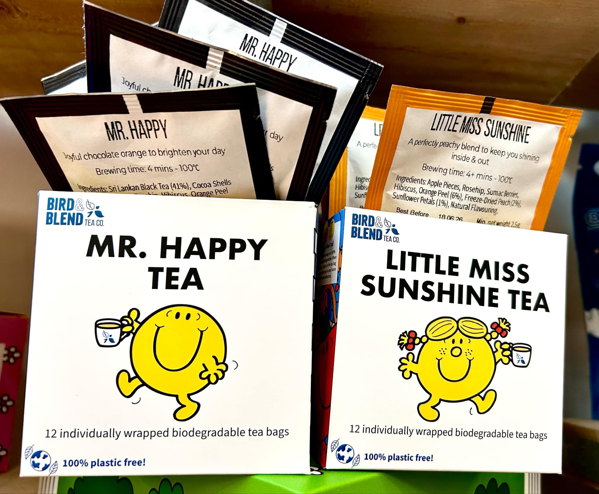 Tea Tastes Terrific | Bird & Blend Tea Company Mr. Men & Little Miss Collection A large rectangular bright yellow cardboard box that has Bird & Blend Tea Company Little Miss Sunshine Gift Cube written in bold black writing and a picture of a bright yellow little miss sunshine that has a smiling face and some flowers in her hair on it next to a short cylindrical clear glass mug full of some dark yellow liquid next to a large rectangular bright yellow cardboard box that has Bird & Blend Tea Company Mr. Happy Gift Cube written in bold black writing and a picture of a bright yellow Mr. Happy that has a smiling face on it next to a short cylindrical clear glass mug full of some dark amber liquid on a light wooden shelf.