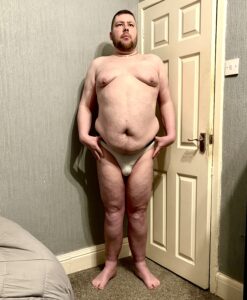 A tall bearded man with his hands on his hips wearing a pair of bright white briefs with a black waistband that has Box Menswear written in bold black writing on it stood against a white wooden door.