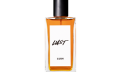 A short rectangular glass perfume bottle full of some translucent red amber liquid with a metallic black lid and a white label that has Lush Lust perfume written in bold black writing on it, on a white background.