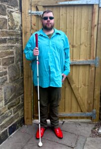 A tall bearded man wearing a pair of silver and black aviator sunglasses with some light green lenses, a light bluey-green long sleeve shirt, a dark khaki green crew -neck t-shirt, a pair of dark khaki green chino trousers and a pair of shiny bright red shoes holding a bright white cane with a red leather handle stood against a dark wooden gate.