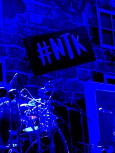 A large bright silver and black drum kit that's illuminated by a bright blue light in front of a dark brick wall that has #NTK written in bold bright blue letters on it, on a dark background.