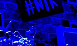 A large bright silver and black drum kit that's illuminated by a bright blue light in front of a dark brick wall that has #NTK written in bold bright blue letters on it, on a dark background.