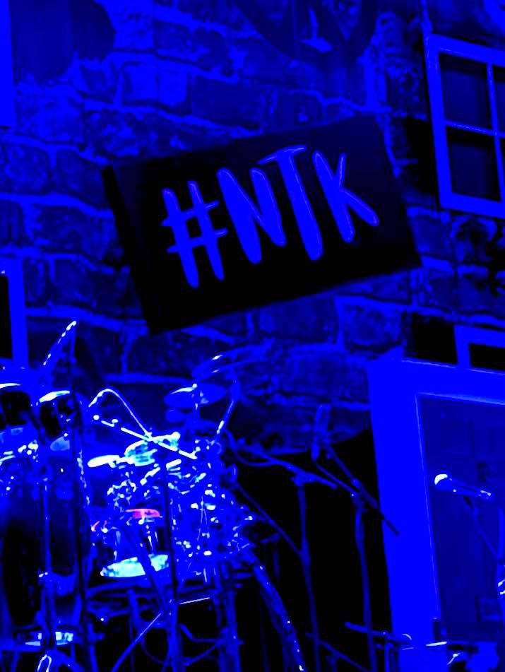 'Cause I'm Not That Kinda Girl | Anastacia Not That Kind Tour York A large bright silver and black drum kit that's illuminated by a bright blue light in front of a dark brick wall that has #NTK written in bold bright blue letters on it, on a dark background.