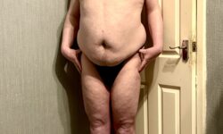 A tall bearded man with his hands on his hips wearing a pair of shiny black tanga briefs with a bright white waistband that has Box Menswear written in bold black writing on it stood against a white wooden door.