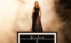 A short Woman with some wavy bright blond hair wearing a stripy Black dress with some sheer panels that reveal her Legs stood on an illuminated stage that’s covered in some bright white lights, has Kylie written in bright white lights on it, and surrounded by some fog, on a white background.