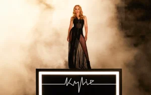 A short Woman with some wavy bright blond hair wearing a stripy Black dress with some sheer panels that reveal her Legs stood on an illuminated stage that’s covered in some bright white lights, has Kylie written in bright white lights on it, and surrounded by some fog, on a white background.